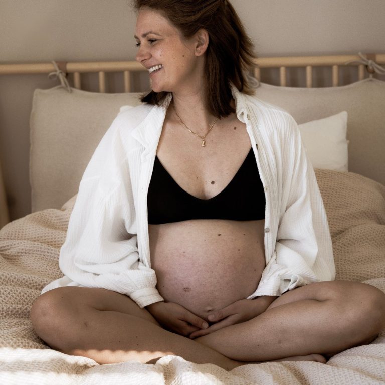Schwangere Frau sitzt lächelnd auf einem Bett, umgeben von weichen Kissen.