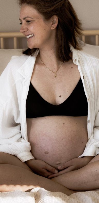 Schwangere Frau mit lächelndem Gesicht sitzt auf einem Bett, bekleidet mit schwarzem Oberteil.
