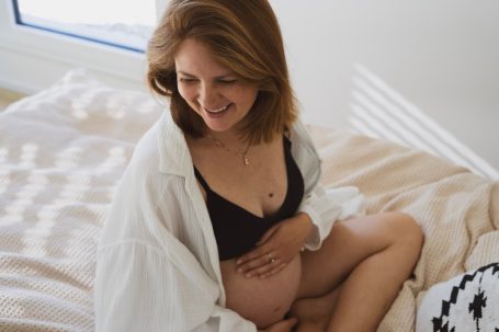 Lachende schwangere Frau in einem weißen Hemd, die ihren Bauch hält.
