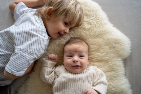 Ein älteres Kind und ein Baby liegen auf einem flauschigen Teppich. Beide lächeln.