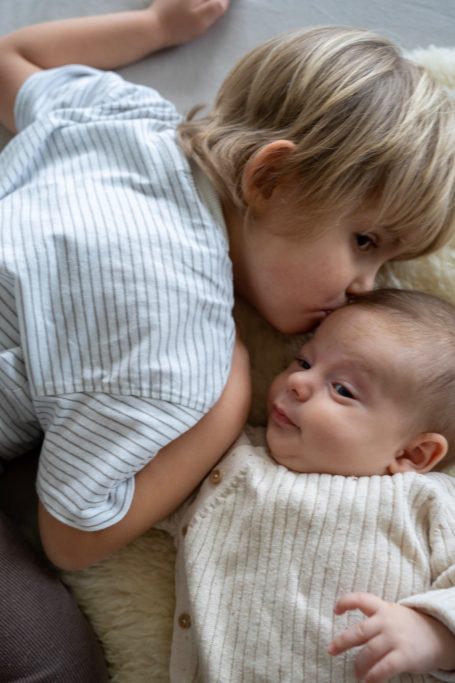 Ein älteres Kind küsst sanft ein Baby, beide liegen auf einem weichen Untergrund.