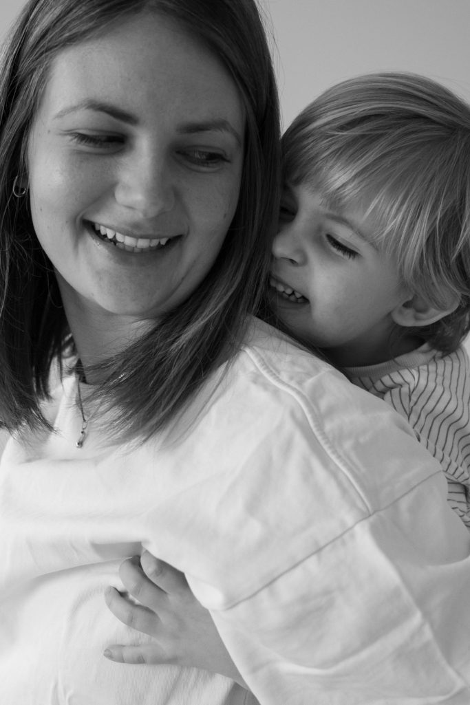Frau mit langen Haaren lächelt, während ein kleines Kind ihr von hinten umarmt.