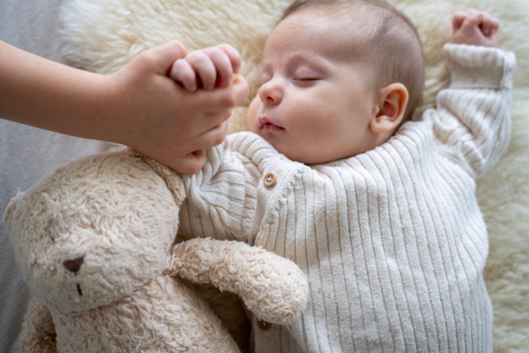 Schlafendes Baby in heller Strickjacke, hält die Hand eines Kindes, umgeben von einem Plüschtier.