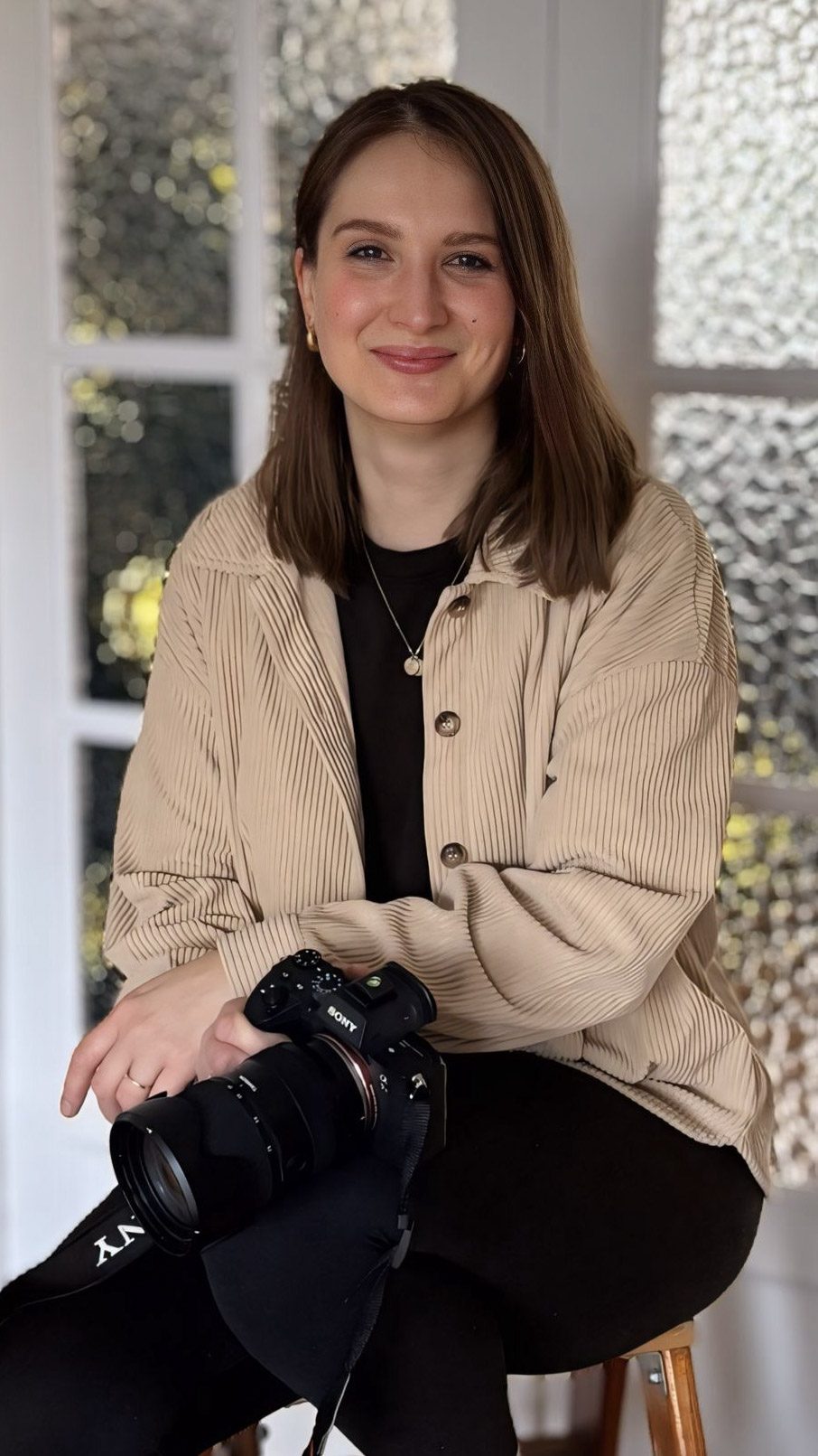 Junge Frau mit Kamera in einer hellen Umgebung, lächelt entspannt in die Kamera.